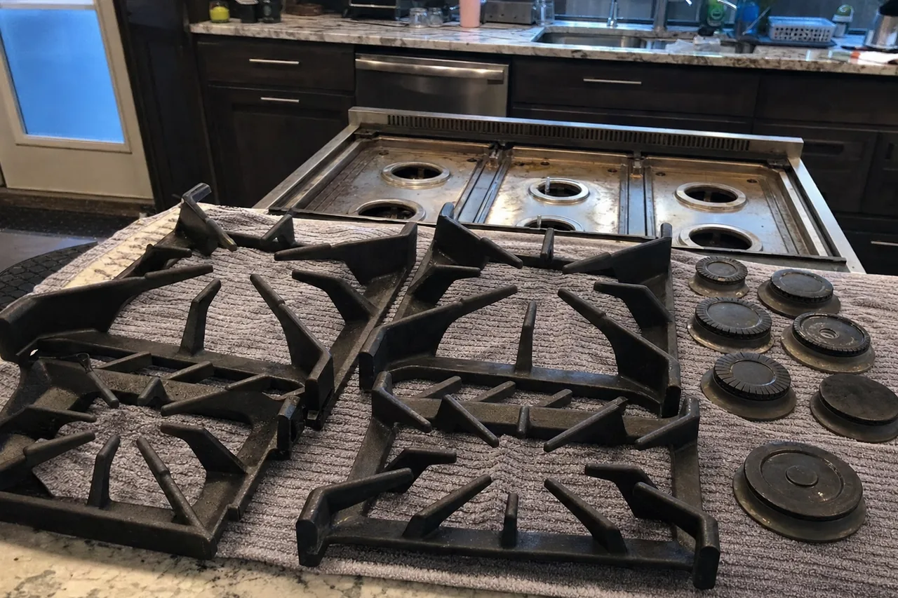 Two cast grate sets and a row of burner caps laid out on a towel on a granite island, a stripped rangetop behind